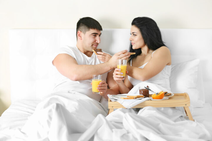 رجل وامرأة يجلسان على السرير وهما يتناولان الطعام معًا في جو حميمي، يرمز إلى الترابط والشراكة والعناية بالصحة الجنسية من خلال نمط حياة متوازن يشمل الغذاء والمكملات الطبيعية.